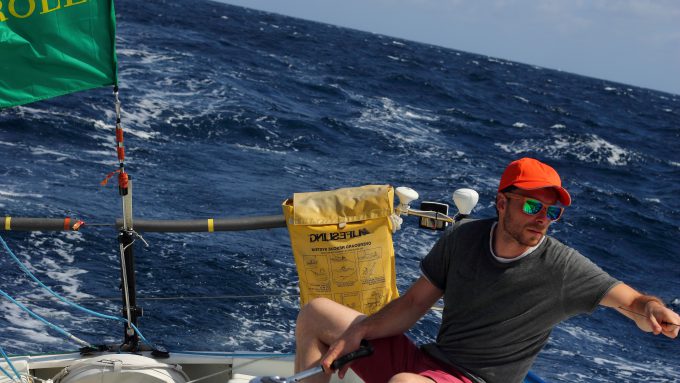 A person in a gray shirt and red hat is sailing in deep blue waters, adjusting a winch handle while pointing with their right hand. A bright green flag and yellow safety equipment are visible on the boat.